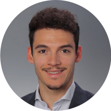 Ein junger Mann mit kurzen lockigen braunen Haaren, heller Haut und einem gestutzten Ziegenbart lächelt in die Kamera. Er trägt ein helles Hemd und einen dunklen Blazer vor einem schlichten grauen Hintergrund.
